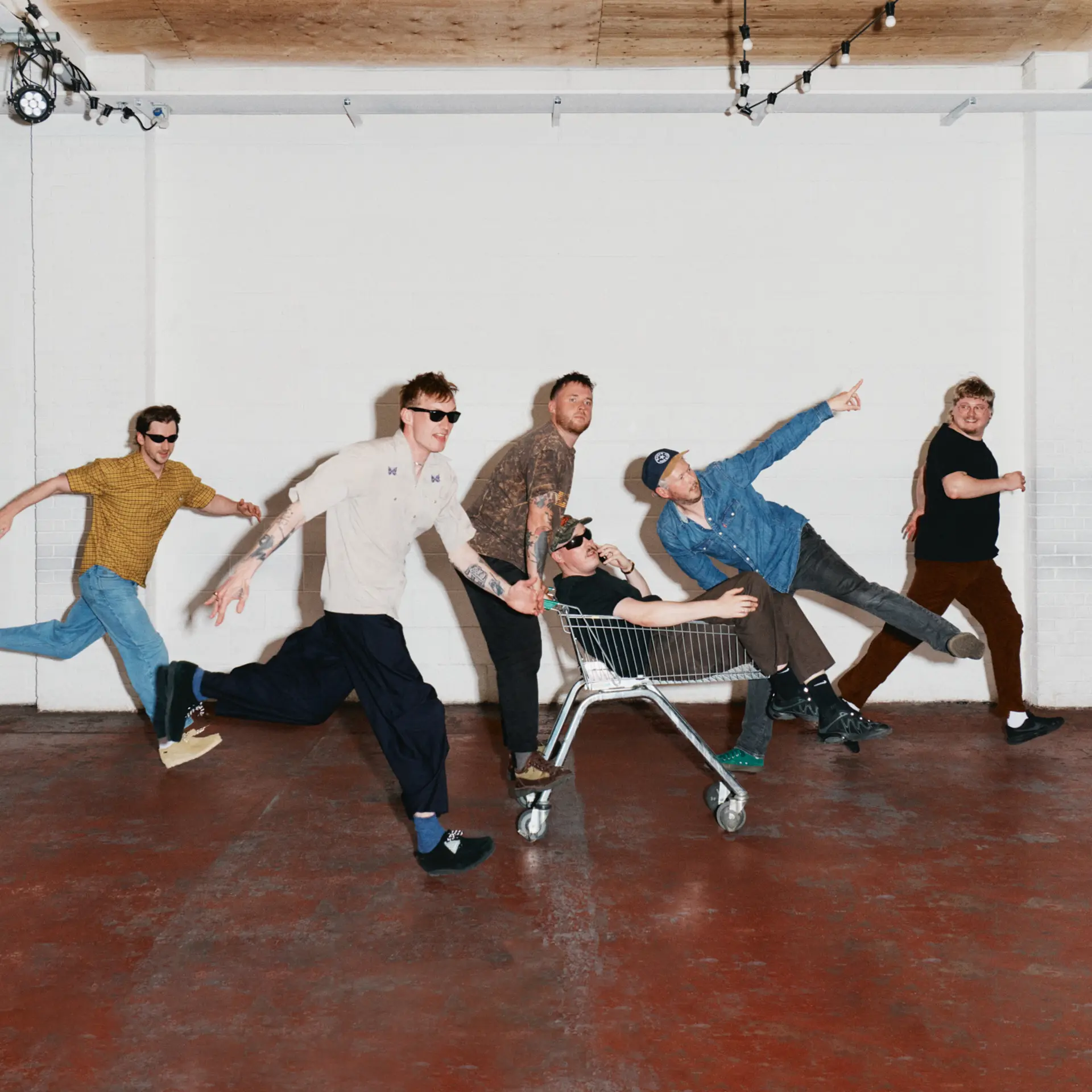 Adult DVD's six members running across a white studio, one riding in a shopping trolley